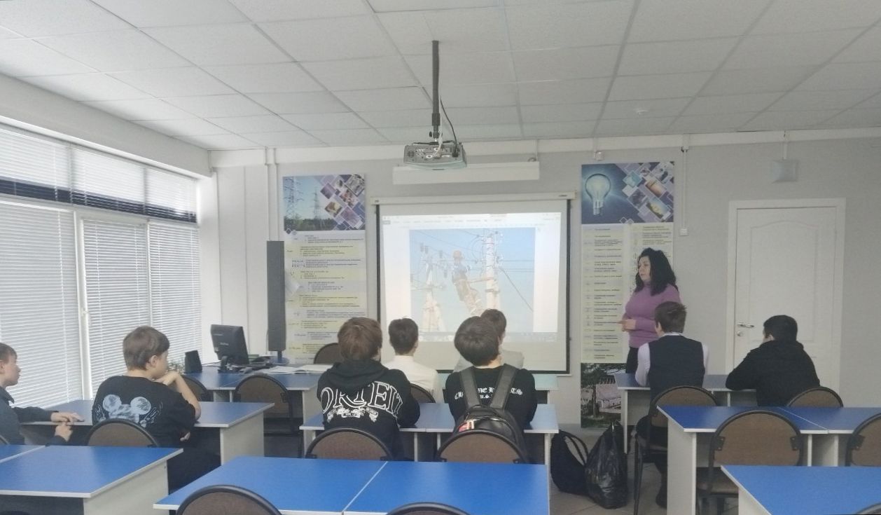 ученики сидят за партами в классной комнате. Они обращены к преподавателю, который стоит у доски с проектором. На экране отображается мультимедийное изображение, вероятно, связанное с темой занятия. В классе много света, и стены оформлены образовательными материалами. Учащиеся, одетые в повседневную одежду, смотрят с интересом на экран и преподавателя.