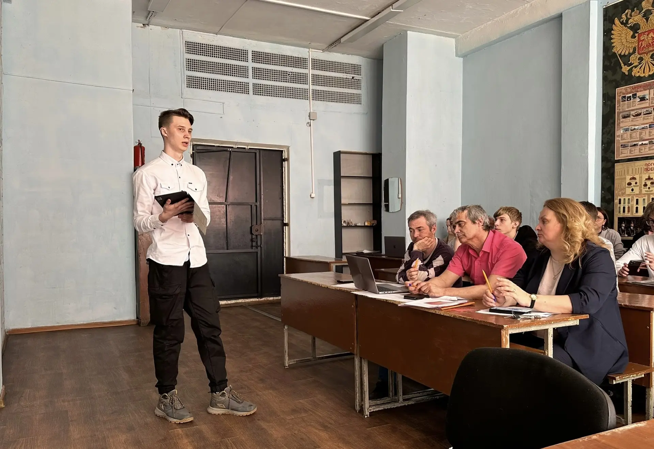 Молодой мужчина стоит у стены в классной комнате или небольшом зале, держит в руках открытый блокнот или папку и говорит перед аудиторией. Он в светлой рубашке и тёмных брюках, на ногах кроссовки. Справа от него длинный деревянный стол, за которым сидят четыре человека — три мужчины и одна женщина — внимательно смотрят на выступающего, у некоторых перед собой ноутбук и бумаги. Помещение с бледными стенами, за спиной видна большая тёмная дверь и пожарный кран.