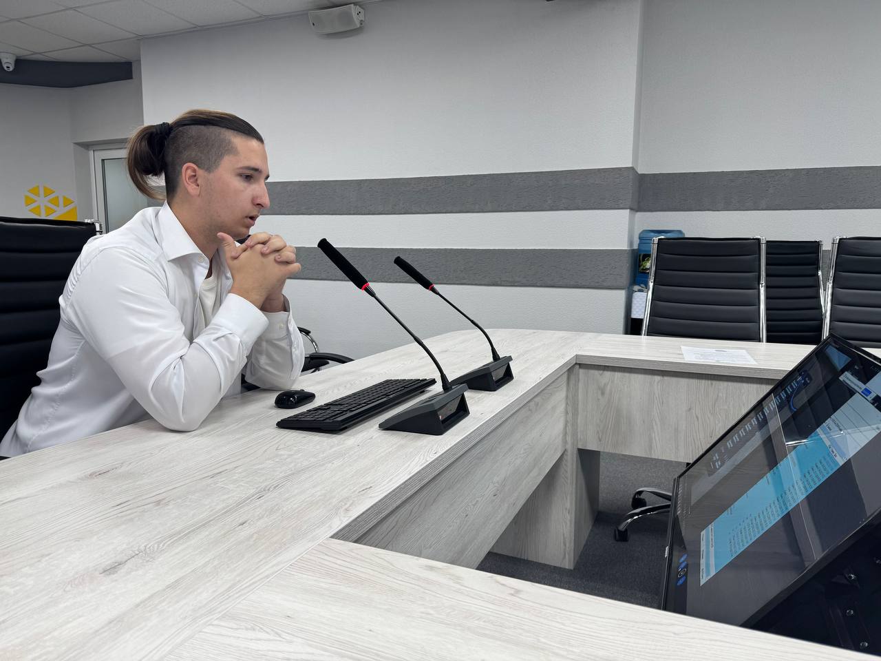 представлен молодой человек, сидящий за столом в конференц-зале. Он одет в белую рубашку, волосы у него длинные и собраны в хвост. Его руки сложены в молитвенной позе на столе, а взгляд устремлен перед собой, где находится экран. На столе — клавиатура и другие предметы, планирующие на встрече. Стены также оформлены в серых и белых тонах, как на предыдущих фотографиях.