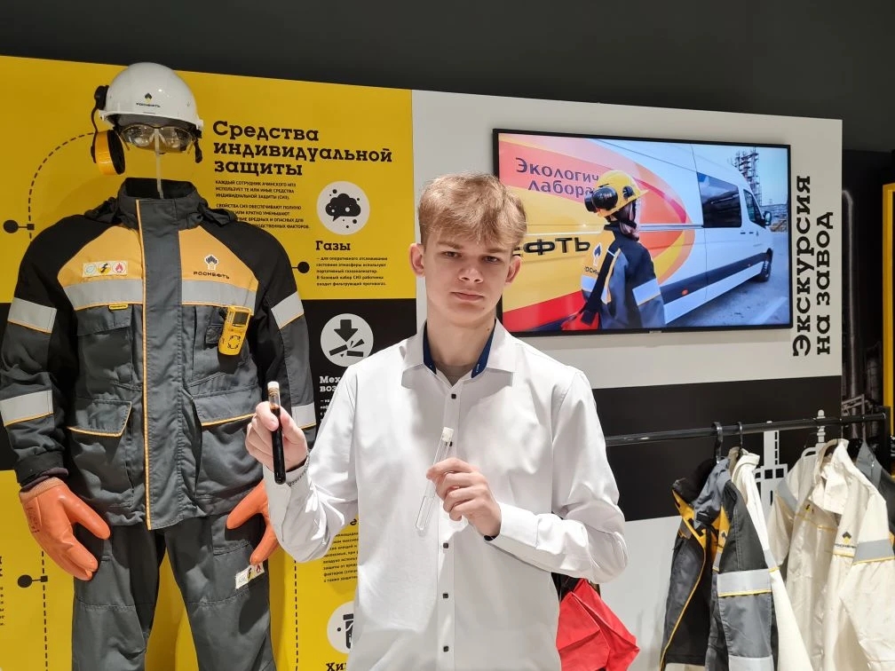 На этой фотографии молодой парень с короткими светлыми волосами одет в белую рубашку и держит в руках небольшой предмет, вероятно, устройство или аксессуар. На заднем плане манекен в рабочей одежде, на стене размещены информационные постеры. Манекен также одет в защитное снаряжение, и вокруг него отображается информация о мерах безопасности.
