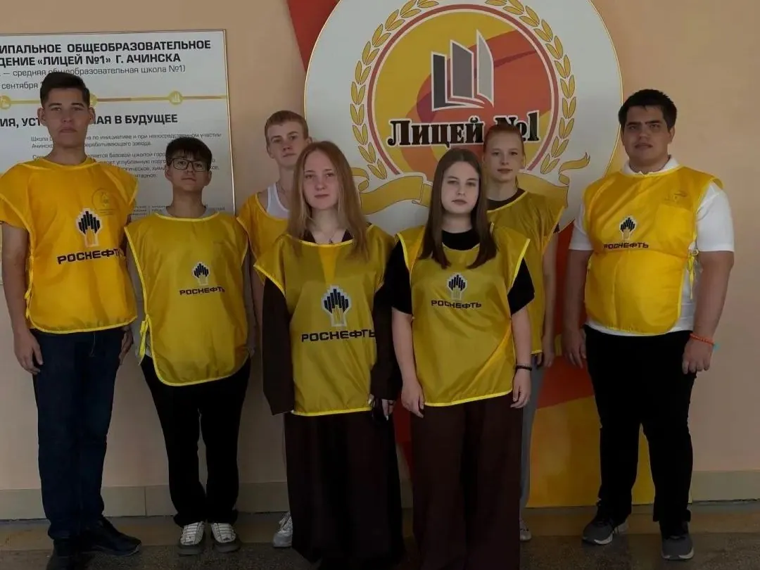 Группа из семи подростков стоит в ряд на фоне баннера «Лицей №1». На школьниках надеты желтые жилеты с логотипом компании «Роснефть». На стене слева расположена информационная табличка с названием учебного заведения.