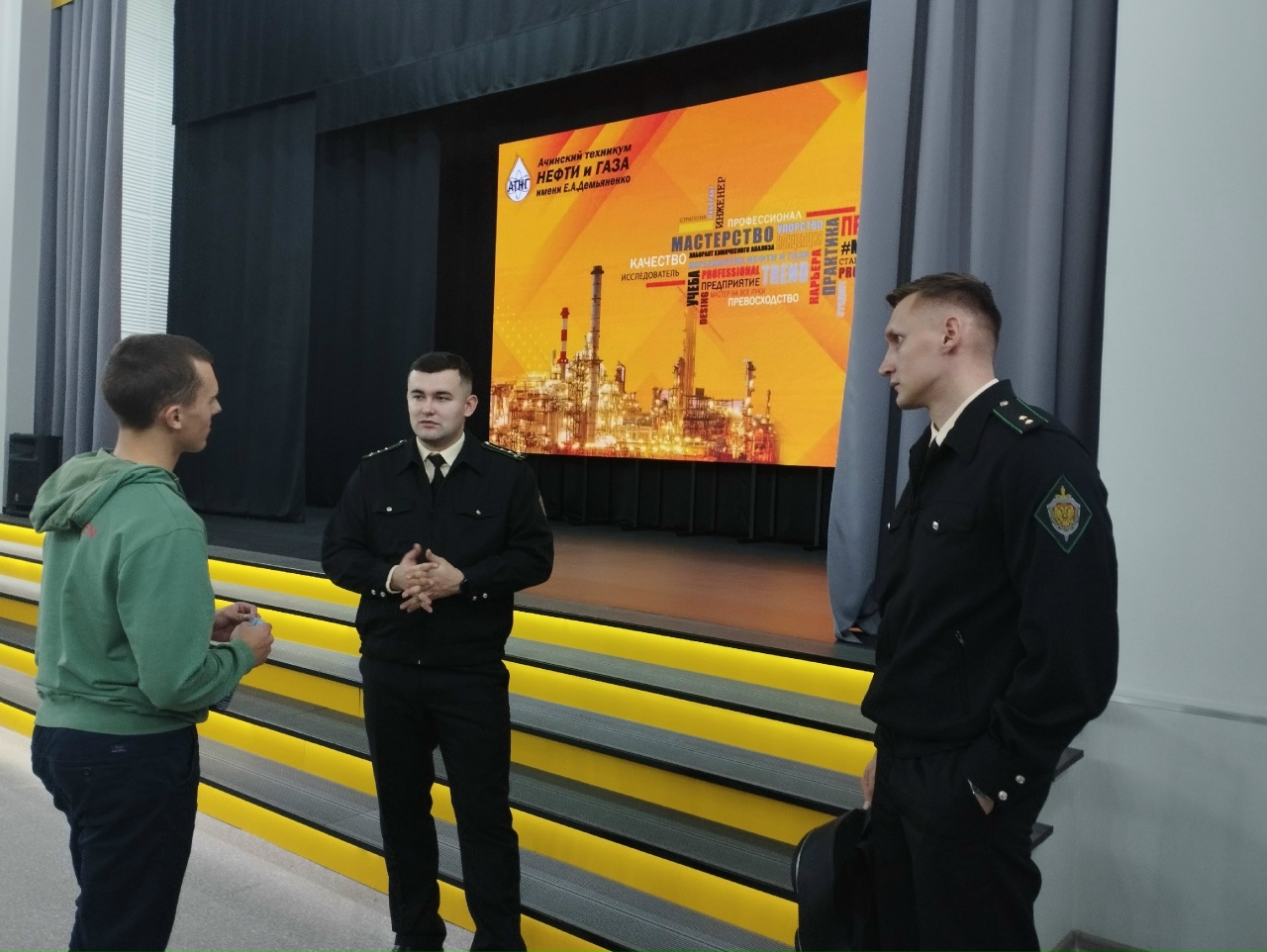 присутствует группа людей, общающихся в помещении. В центре - молодой человек в зеленом худи, который разговаривает с двумя мужчинами в форменной одежде. Мужчины выглядят серьезно и внимают собеседнику. На заднем плане находится большая сцена с ярким экраном, на котором изображены тематические графические элементы, возможно, связанные с промышленностью.