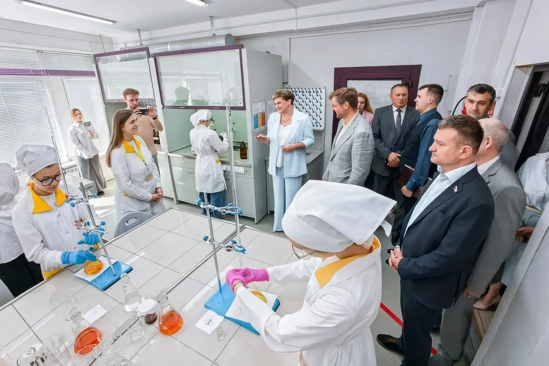 Делегация чиновников наблюдает за экспериментом в химической лаборатории. Дети-лаборанты в белых халатах, защитных очках и перчатках работают с колбами и штативами на лабораторных столах.