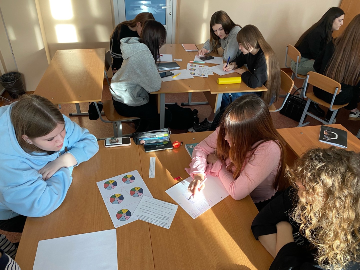 изображена группа из восьми девушек, собравшихся за столом в светлом помещении. Они активно занимаются чем-то, записывают и общаются между собой. Некоторые из них смотрят на свои телефоны, в то время как одна девушка с длинными волосами в центре рисует или делает заметки на белом листе, окруженном яркими цветными кругами. Солнечный свет освещает стол, создавая теплую атмосферу.