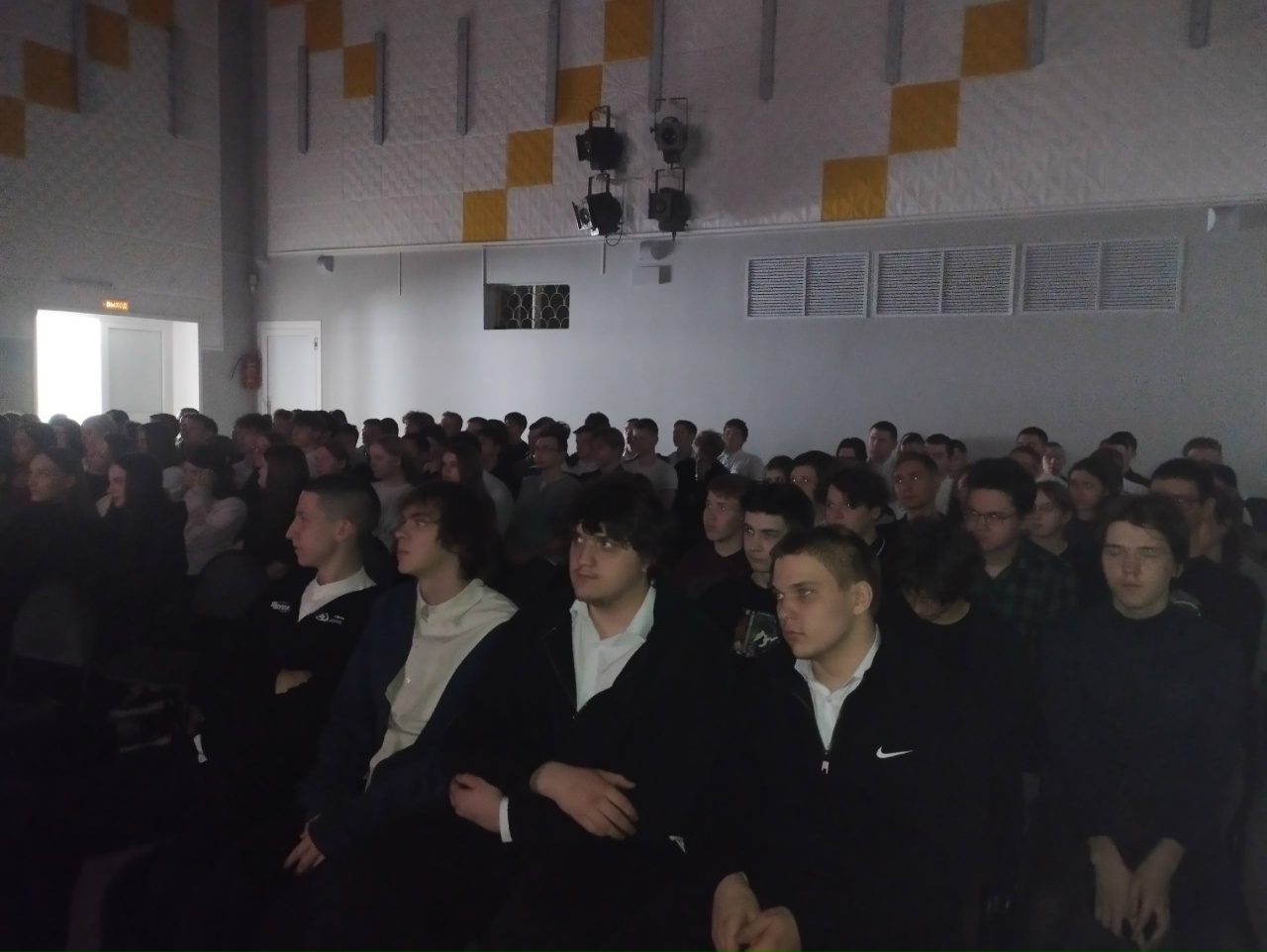 изображена большая группа людей, сидящих в зале. В центре кадра большинство зрителей, одетых в светлые и темные рубашки, сосредоточены на какой-то презентации или мероприятии. Зал оформлен в светлых тонах с яркими элементами, которые выделяются на фоне. На стенах видны оконные проемы и дайте вентиляции.