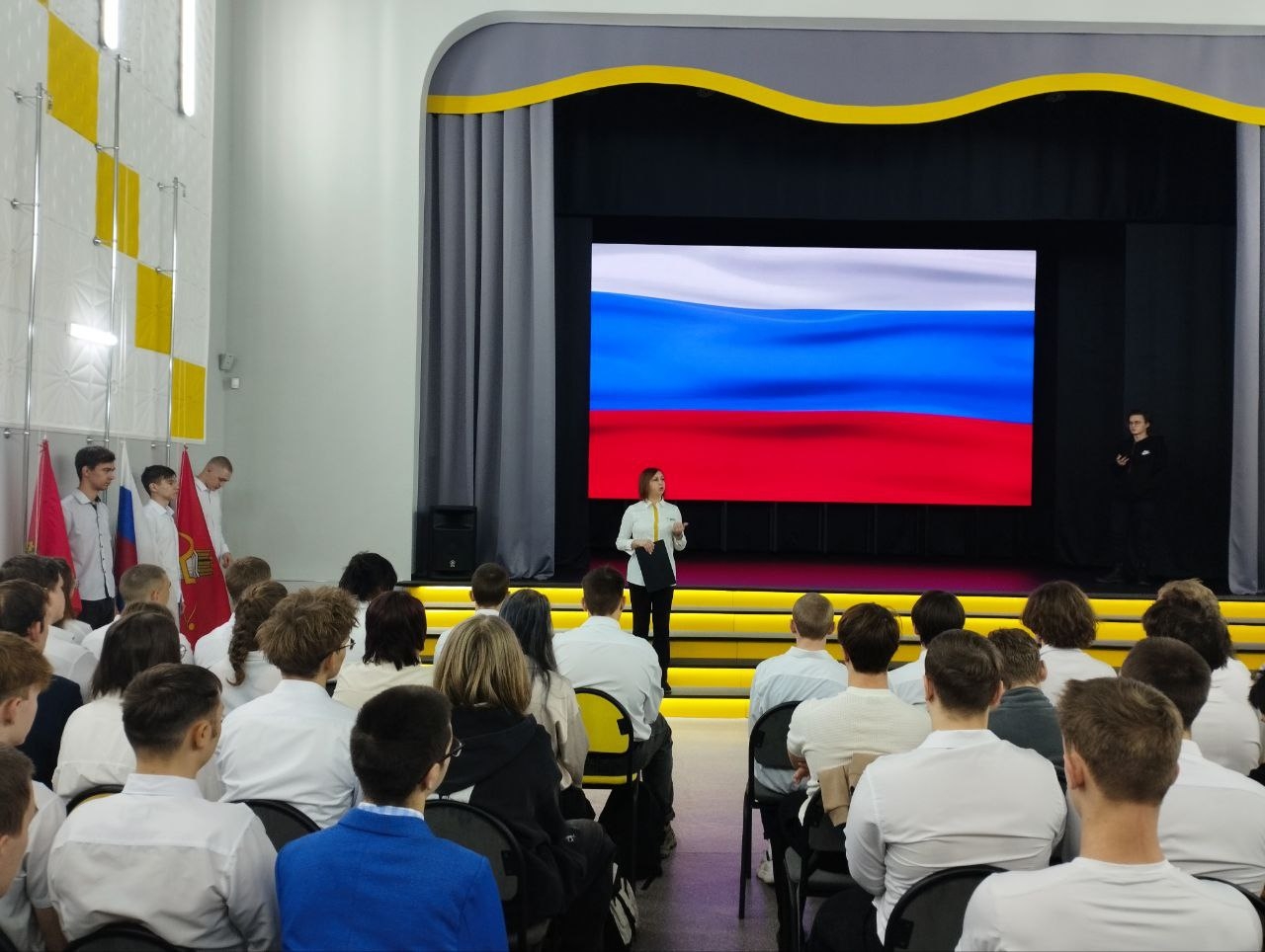 На этом фото видно большое помещение, в котором собралось множество людей. На сцене стоит женщина, выступающая с речью, позади нее на большом экране изображен триколор России (белый, синий, красный). По бокам сцены находятся те же флаги, что и на первом фото. Зрители, сидящие в аудитории, одеты в светлые рубашки и внимательно смотрят на сцену.