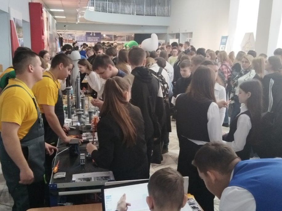 На этом фото изображено большое количество молодых людей, собравшихся в помещении. В центре кадра видны столы, за которыми работают группы студентов. Один из студентов в яркой желтой футболке обслуживает посетителей. Люди активно общаются между собой, исследуя различные проекты и экспериментальные установки. Фон заполняют декорации и плакаты с информацией о мероприятии, а также цветные шарики.