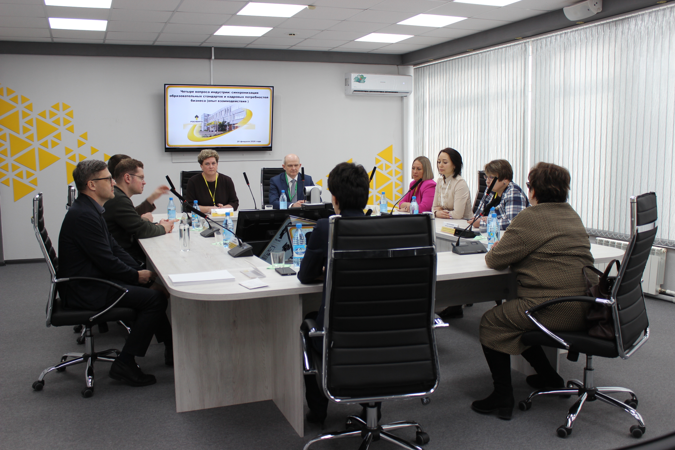 На фото изображена современная конференц-комната. В центре стола находятся несколько человек, сидящих в креслах. Участники обсуждения выглядят сосредоточенными и вовлеченными в разговор. На стене висит экран с текстом или презентацией. Комната оформлена в светлых тонах, с большими окнами, что обеспечивает хорошее освещение. По периметру стола стоят бутылки с водой и цветные папки. В основном, участники выглядят в деловой одежде, что подчеркивает официальный характер встречи.