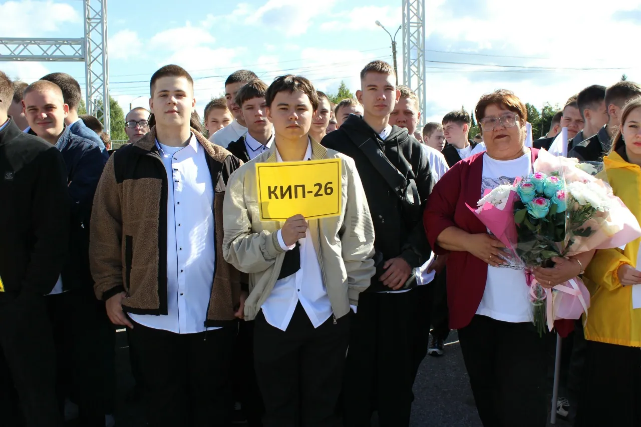 На улице на фоне открытого пространства собралась группа студентов. В центре молодой человек держит желтую табличку с надписью «КИП-26». Рядом с ним девушка с большим букетом цветов, вокруг — другие студенты, ожидающие начала или завершения мероприятия.