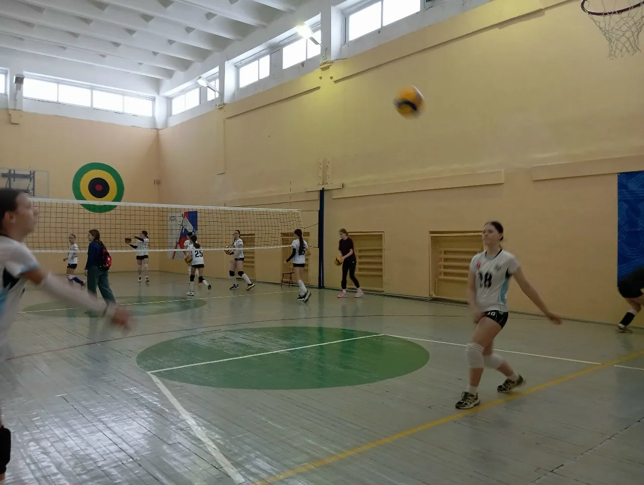 Девушки играют в волейбол в просторном спортивном зале с желтыми стенами и высокими окнами. На деревянном полу с зелеными зонами разметки. В воздухе справа летит желто-синий мяч. На переднем плане справа девушка в белой футболке с номером 18, черных шортах и наколенниках готовится принять подачу. Слева видна размытая фигура игрока в движении. За сеткой на заднем плане находятся другие участницы и тренер. На стене висит мишень для стрельбы из лука, а справа вверху — баскетбольное кольцо.