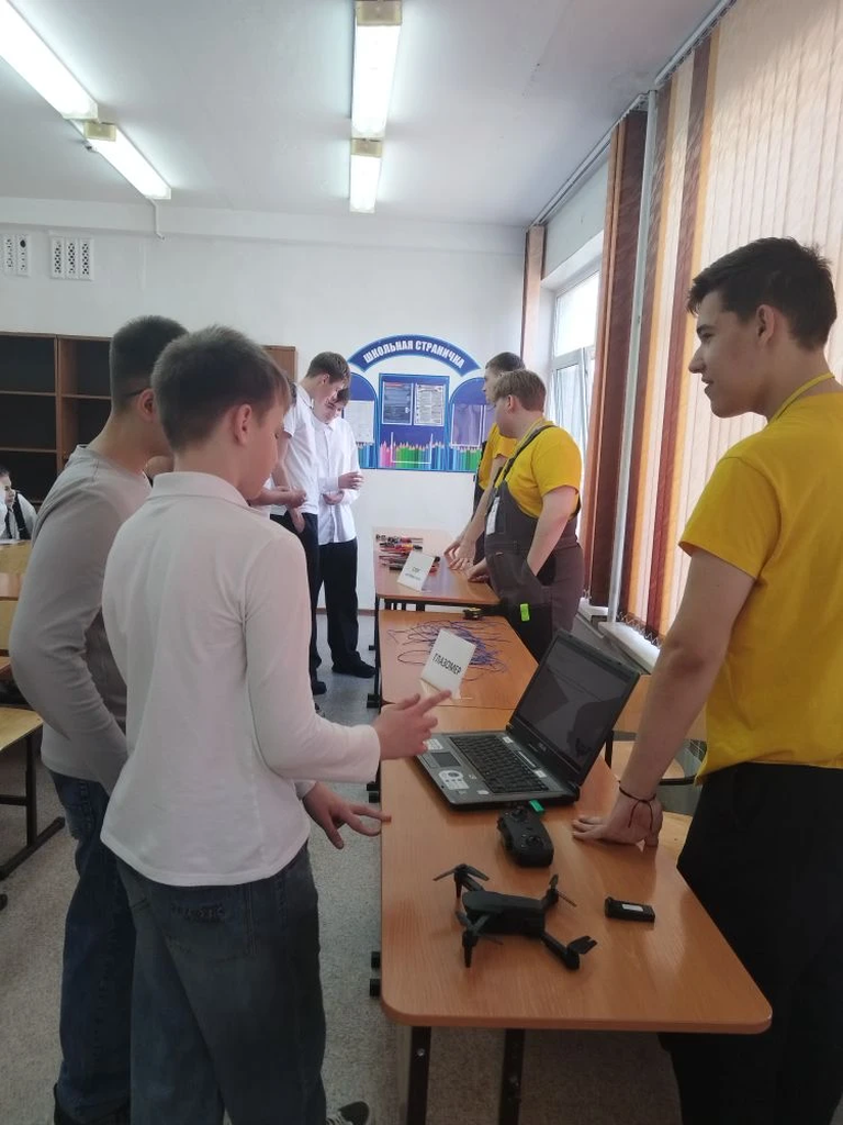 группа подростков взаимодействует с обучающими ресурсами. Два юношы в желтых футболках стоят за столом, на котором расположены компьютерные устройства и один из них держит дрон. Учащиеся внимательно слушают, что им объясняют.