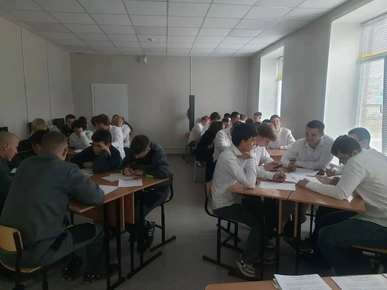 В светлой учебной аудитории с потолочными лампами и окном с жалюзи за столами сидят молодые люди. Часть из них в серых рабочих комбинезонах с желтыми светоотражающими полосами, часть — в белых рубашках. Они пишут на листах бумаги, некоторые разговаривают. На столах лежат бумаги и ручки. Вдалеке видна дверь и стена.