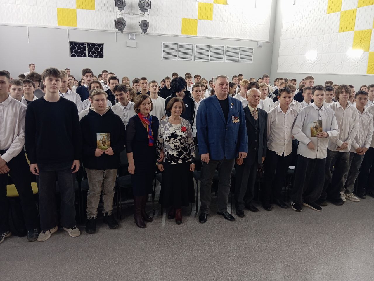 группу людей, стоящих на сцене. В центре – женщина в белой блузке с цветочным принтом, рядом с ней мужчина в синем пиджаке, а также несколько других людей, среди которых заметны пожилой мужчина в темном пиджаке и молодые мужчины в белых рубашках. Все они позируют для фотографии, держа в руках книги. Сзади – группа молодых людей, как правило, в однородной одежде, смотрящих на камеру. Зал оформлен в современном стиле с жёлтыми элементами декора.