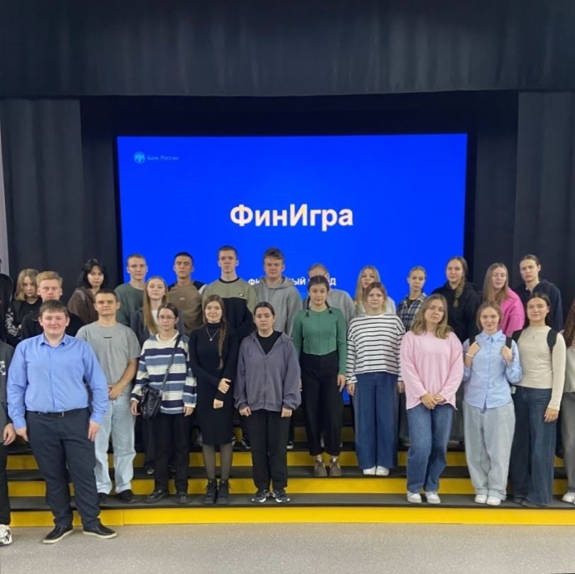 На этой фотографии видно 27 человек, стоящих на сцене. Все участники расположены в несколько рядов, и они выглядят радостными. Задний фон большой и темный, с ярким экраном, на котором написано слово "ФинИгра". Участники одеты в повседневную одежду, каждое лицо излучает уверенность и позитив.