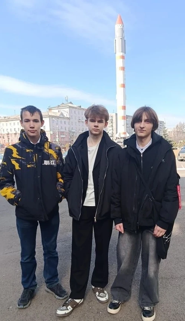 Три подростка стоят в ряд на открытом пространстве на улице, позади них — высокая декоративная ракета на пьедестале и городские здания. Все трое в тёмных куртках: слева — тёмная куртка с жёлтыми пятнами и джинсами, в центре — чёрная тонкая куртка поверх светлой футболки и свободные чёрные штаны, справа — тёмная куртка и серые свободные штаны. У всех серьёзные выражения лиц, держатся близко друг к другу; день солнечный, небо голубое.