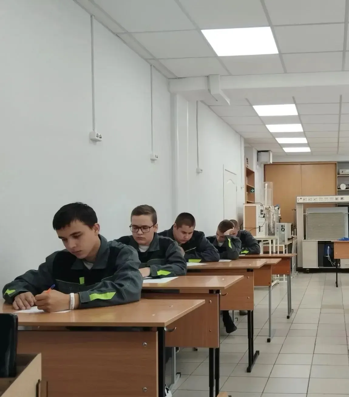 Светлая учебная аудитория с длинными столами, за которыми сидят студенты. Молодые люди одеты в одинаковые серые рабочие комбинезоны с желтыми светоотражающими полосами. На переднем плане студент пишет ручкой в тетради, рядом с ним еще несколько человек также заняты письмом. В глубине класса видно лабораторное оборудование и шкафы. Освещение яркое, потолок с квадратными светильниками.