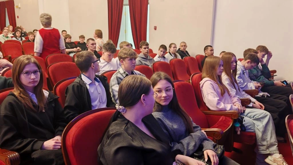Интерьер театрального зрительного зала с красными плюшевыми креслами; в первых рядах сидит группа подростков и молодых людей в повседневной одежде — джемперы, рубашки и светлые джинсы. Некоторые повернуты друг к другу и разговаривают, одна девушка смотрит прямо в камеру с лёгкой улыбкой; позади них виднеются занавески и стены светлого тона. Свет ровный, дневной; обстановка спокойная, ожидание начала спектакля.