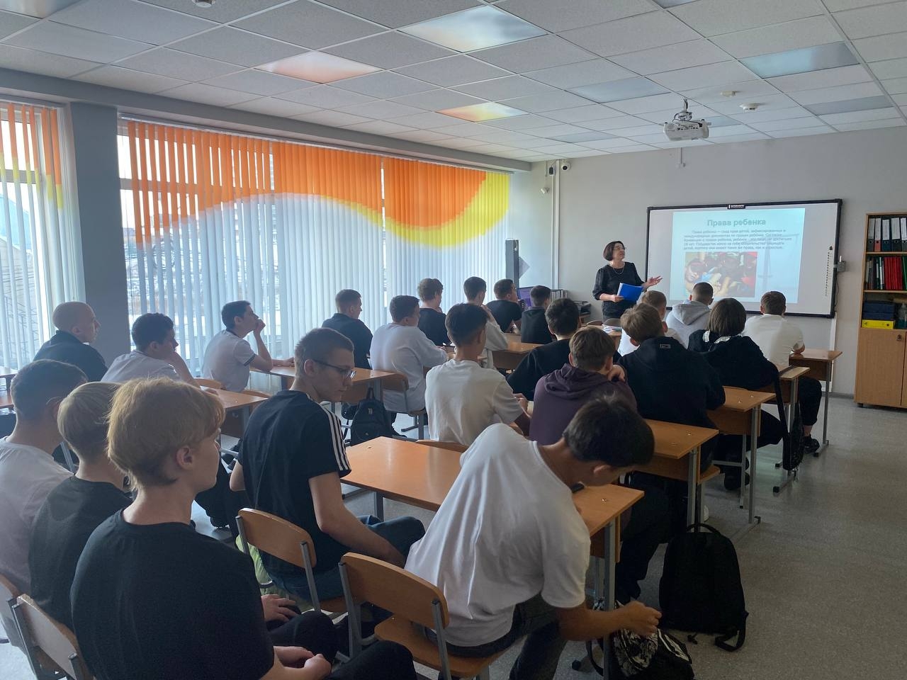 показывает классную комнату, где проходит урок или презентация. В центре кадра стоит преподаватель с документами, обращённый к группе из около двадцати учеников, сидящих за партах. На стенах видны большие окна с яркими шторами, а на стенде расположены учебные материалы. Большинство учащихся одеты в формальную или полуформальную одежду. Урок выглядит увлекательным, а внимание студентов сосредоточено на лекторе.