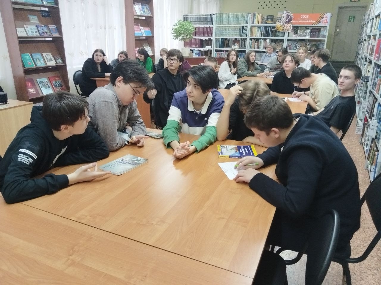 На этом фото несколько студентов активно общаются за столом. Они обсуждают темы, делятся идеями и, возможно, готовятся к презентации или заданию. У одного из мальчиков в руках книга, что указывает на связь с учебным процессом. Позади них находятся другие студенты, что создает динамичную атмосферу общения и сотрудничества. Библиотека снова служит фоном, указывая на окружение, способствующее обучению.