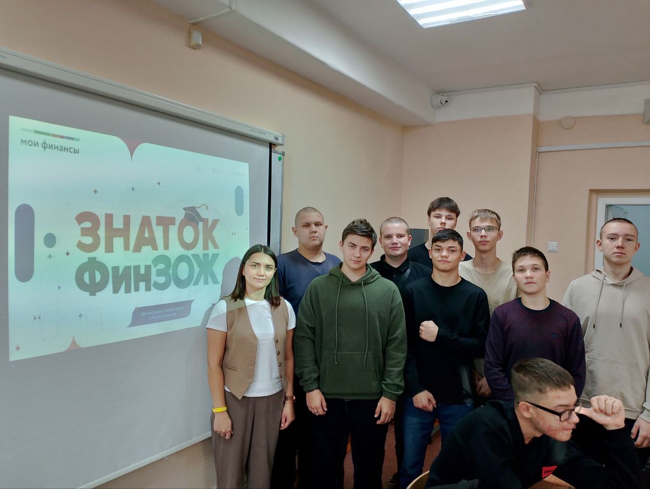На третьем фото показана группа молодых людей, стоящих перед слайдом на стене с изображением заголовка «ЗНАТОК ФИНЗОЖ». Все участники смотрят в камеру. У каждого из них неформальная одежда, а на лицах — заинтересованные выражения. Одна девушка стоит в центре, а остальные стоят и сидят вокруг неё.