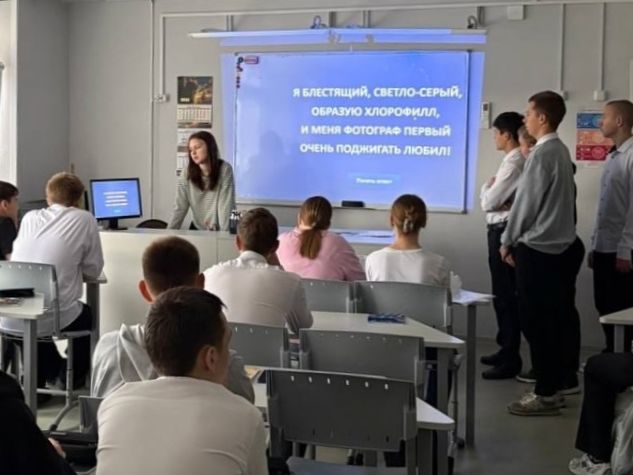В классе, где ученики сидят за партами, а на экране проектора отображается текст. Женщина, вероятно, учитель, стоит у доски и объясняет материал. Ученики, одетые в светлую и темную одежду, внимательно слушают. На экране написано: "Я блестящий, светло-серый, обожаю хлорофилл, и меня фотографируют первый раз. Очень поднимать люблю!"
