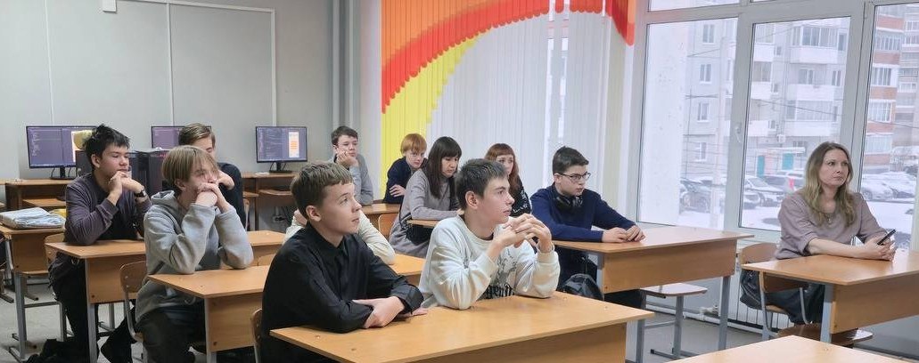 видно группу студентов, сидящих за столами в классе. Они внимательно смотрят вперед, скорее всего на преподавателя или проектор. Учащиеся разнообразно одеты, большинство из них в темных тонах. На заднем плане видны несколько компьютеров, а в окне — вид на жилые дома. Атмосфера сосредоточенная и внимательная, что создает впечатление учебного процесса.