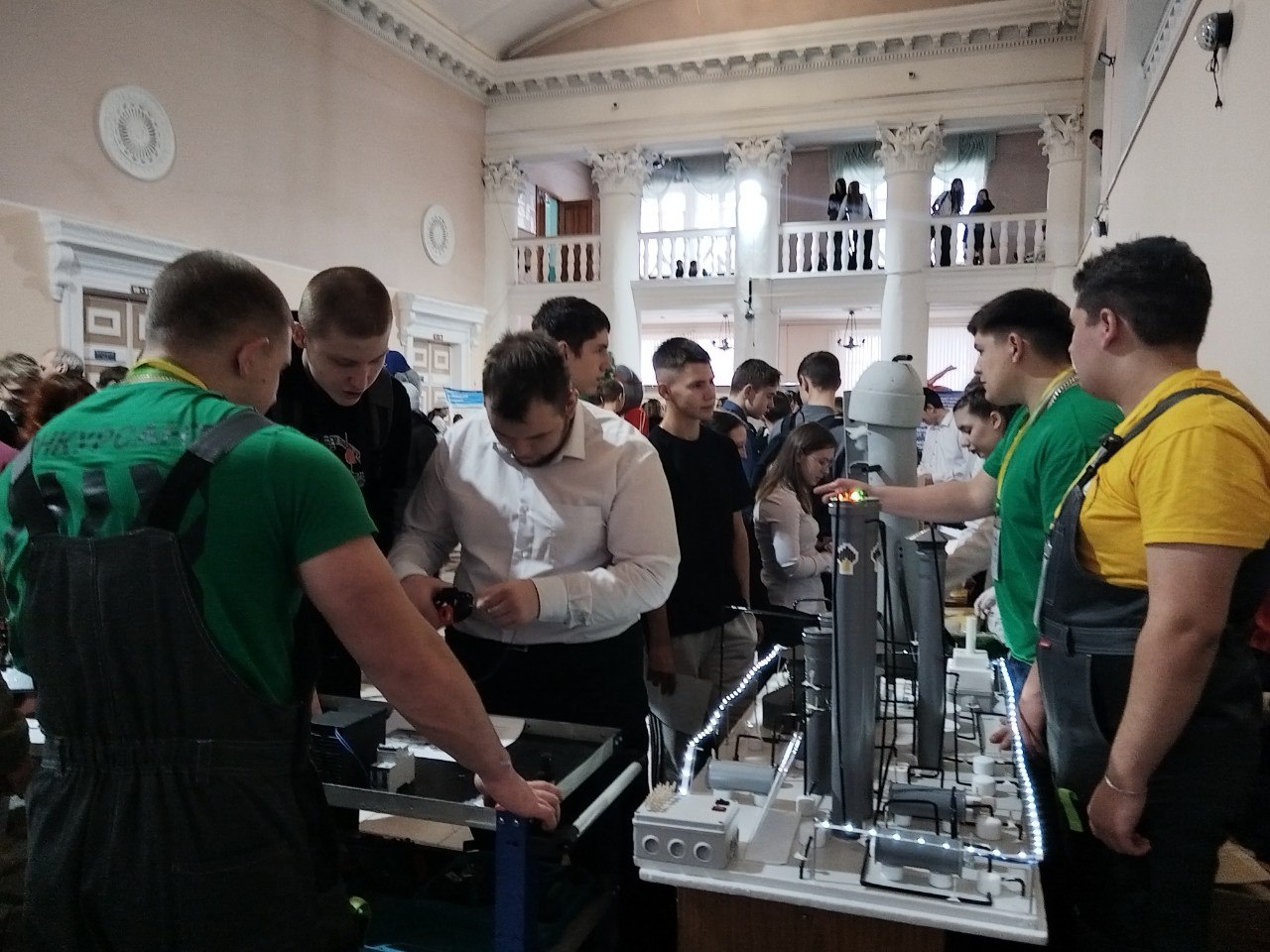 На фотографии изображена группа людей, собравшихся в помещении с высоким потолком и колоннами. По центру находится выставка с моделями, представленными на столах. Некоторые участники активно обсуждают представленный проект, в то время как другие внимательно изучают детали. На столе лежат конструкции, напоминающие элементы техники или робототехники. Видно, что участники одеты в разные цвета униформы: некоторые в зеленых футболках, другие в серых комбинезонах.