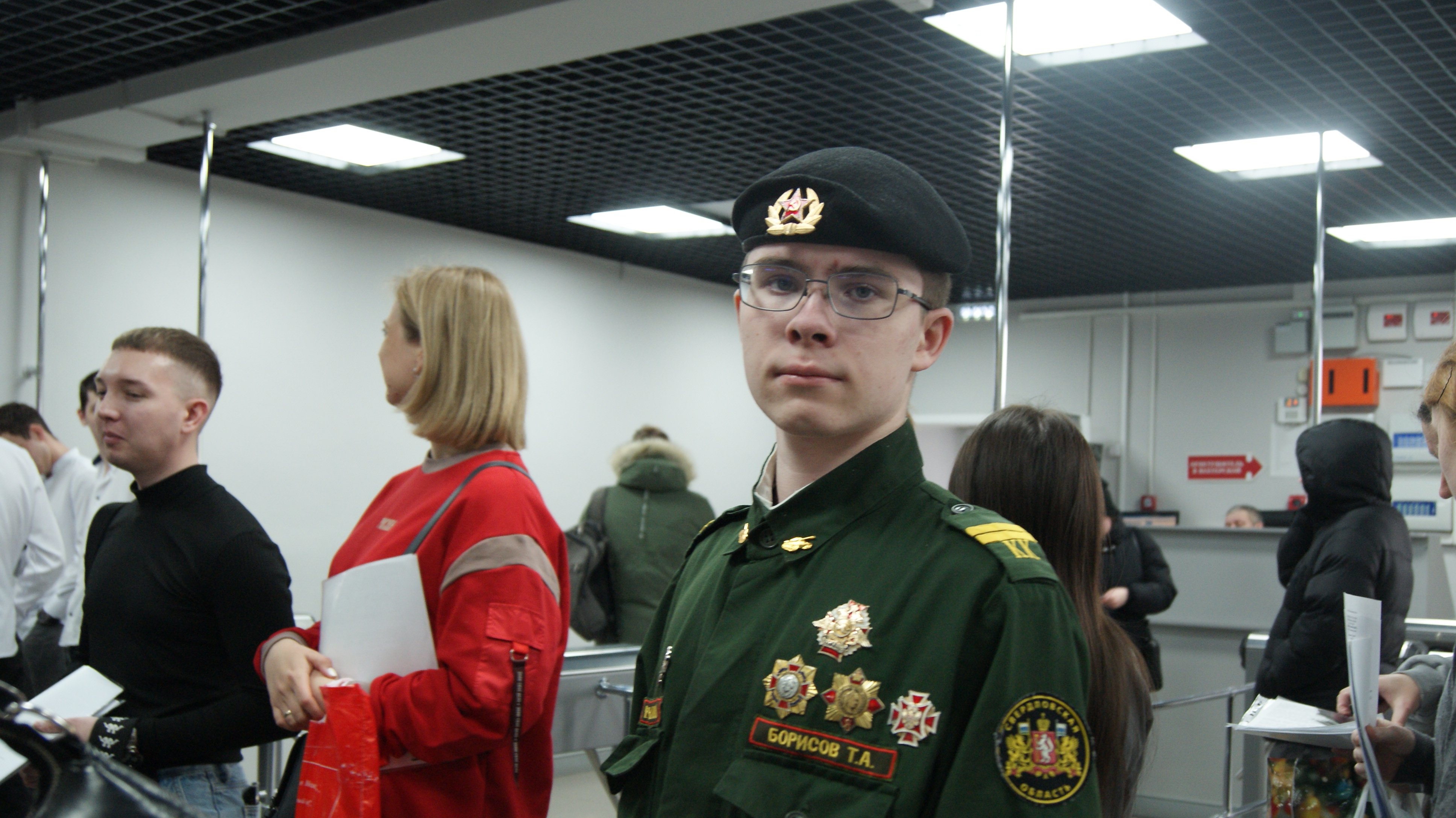 Молодой человек в тёмно-зелёной военной форме и фуражке смотрит в камеру; позади него — несколько стоящих людей в повседневной одежде, потолок с квадратными светильниками и зеркальная стена.