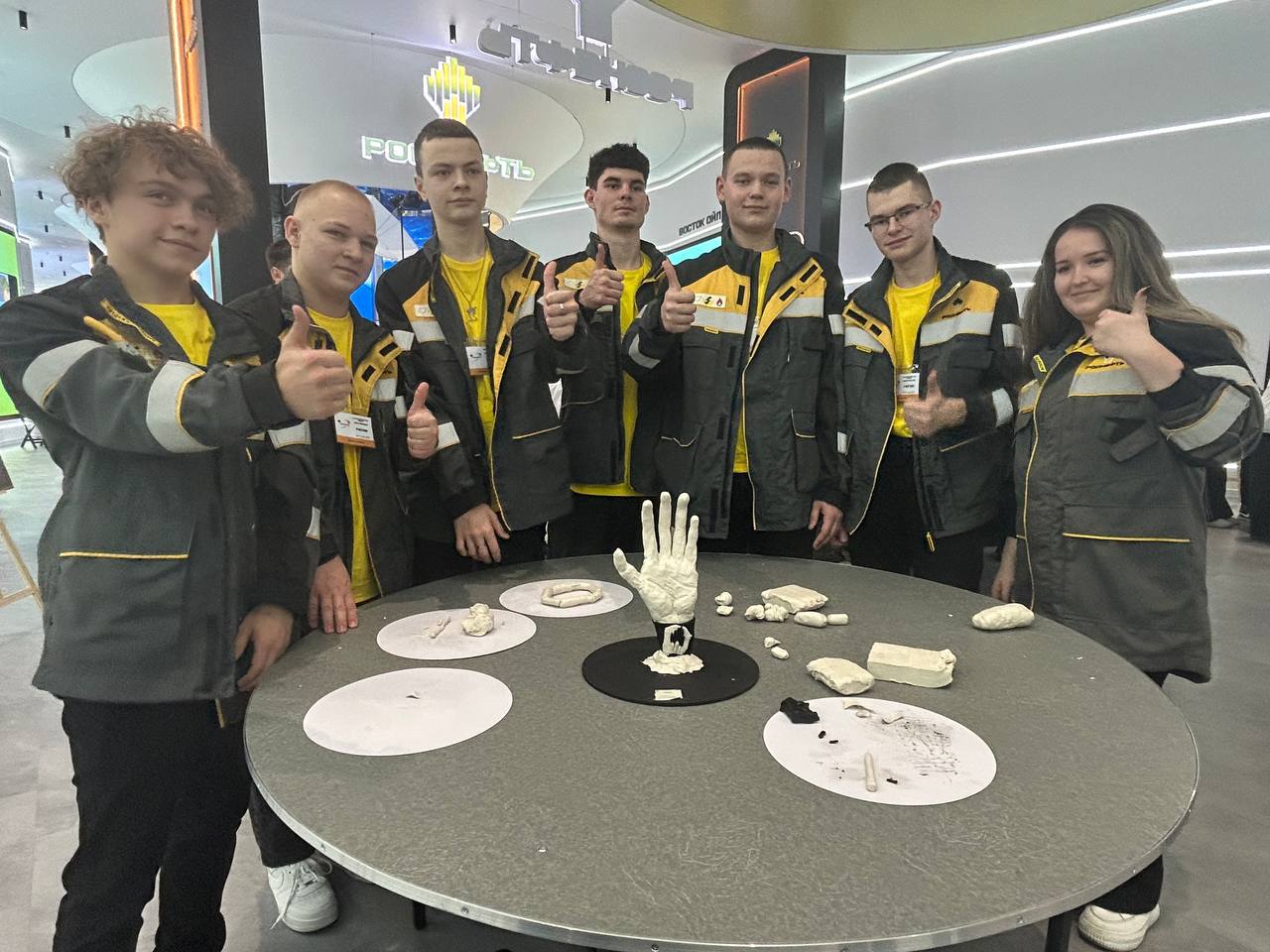 На этом фото группа молодых людей в серых куртках с желтыми акцентами позирует для снимка. Они стоят около круглого стола, на котором размещены предметы, напоминающие гипсовые слепки и другие материалы. Все участники улыбаются и показывают знак "ладонь вверх", выражая позитивные эмоции.