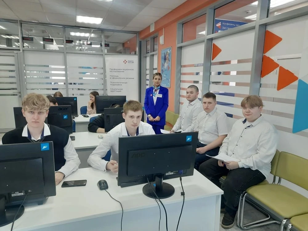 фото молодые люди сидят за компьютерами в офисном помещении. Трое из них смотрят в камеру, а другие находятся за ними. Женщина в официальной одежде стоит позади, наблюдая за процессом. Окружение составляет современное офисное пространство с яркими цветами и хорошим освещением, создающее уютную атмосферу для работы.