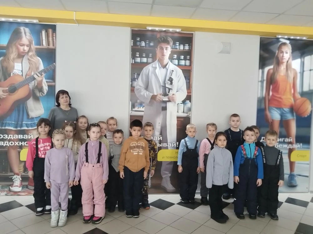 На фотографии изображена группа детей, стоящих в помещении. Позади них размещены большие плакаты с изображениями подростков: одна девушка играет на гитаре, а другой парень в белом халате, видимо, в роли ученого, держит пробирку. Девочка с мячом для баскетбола смотрит в сторону. Дети находятся в центре кадра, одеты в разноцветные спортивные костюмы и комбинезоны. Весьма хорошее освещение.