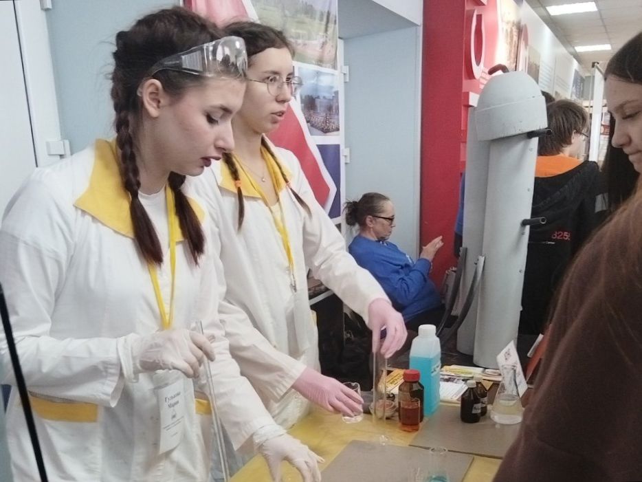 На фотографии показаны два человека, стоящие за столом. Обе девушки одеты в белые халаты и защитные очки, на них есть желтые нагрудники. Одна из них с длинными волосами, заплетенными в косички, а другая — с длинными распущенными волосами. Они занимаются научным экспериментом, на столе размещены разнообразные химические жидкости в прозрачных и темных бутылках. В заднем плане видно людей, сидящих на стульях и наблюдающих за происходящим.