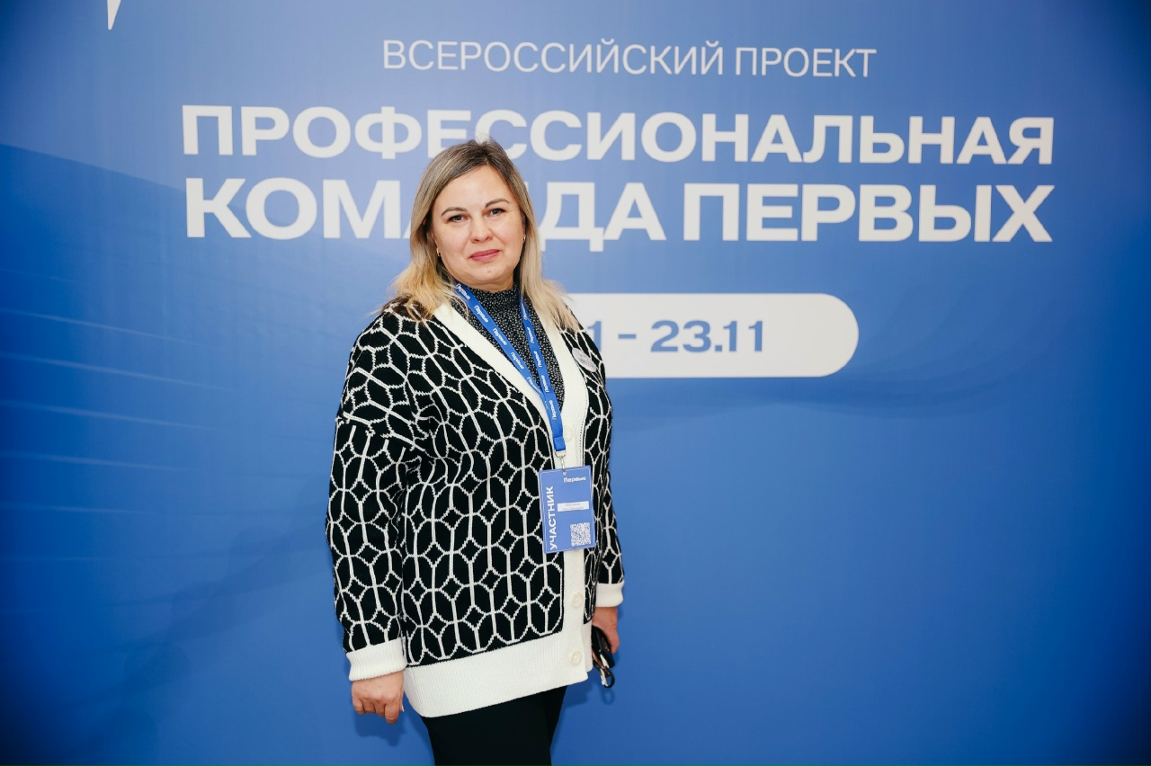 На фото изображена женщина, стоящая на фоне синей стенды с белым текстом. Она одета в черно-белый свитер с геометрическим узором и улыбается. На ней видна бадж с логотипом мероприятия, а также бирюзовый ленточка на шее. Фон стены содержит надпись о всероссийском проекте, который, вероятно, подразумевает профессиональное событие или конференцию.