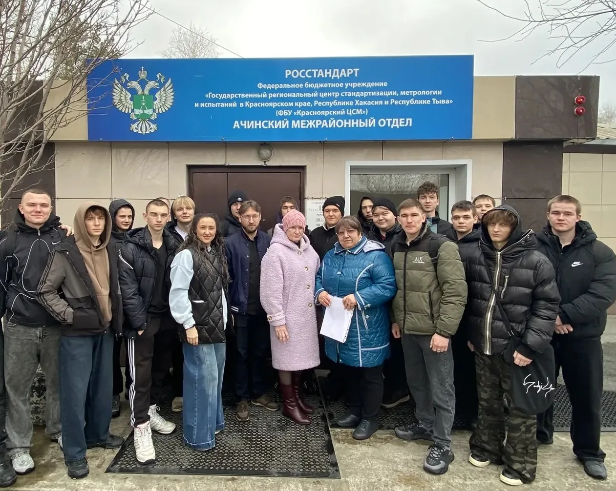 На фото изображена группа из двадцати людей, стоящих перед зданием, обозначенным знаком "Росстандарт". В центре находятся мужчины и женщины, некоторые из которых держат документы. Все участники улыбаются, на них одеты тёплые одежды, так как видны облака на небе и признаки холодной погоды.
