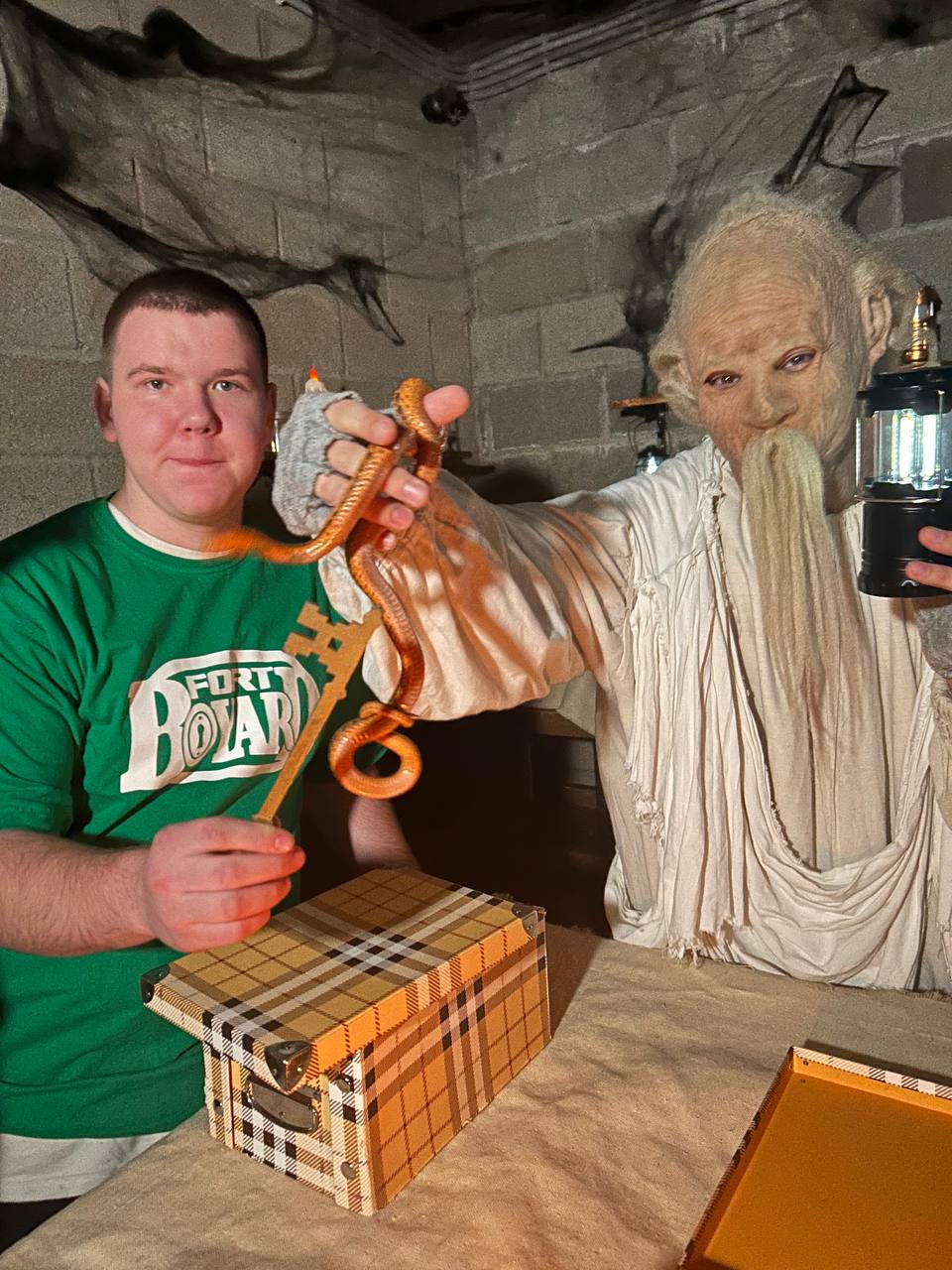 На изображении молодой человек в зеленой футболке держит змею, выставленную вперед. Его левая рука параллельна столу, на котором лежит коробка в клетку. Рядом с ним стоит пожилой мужчина с длинной белой бородой, одетый в светлую одежду. Он держит фонарик и смотрит на камеру с загадочным выражением лица. Фон составляет слегка затемненная комната с элементами декора, создающими атмосферу.