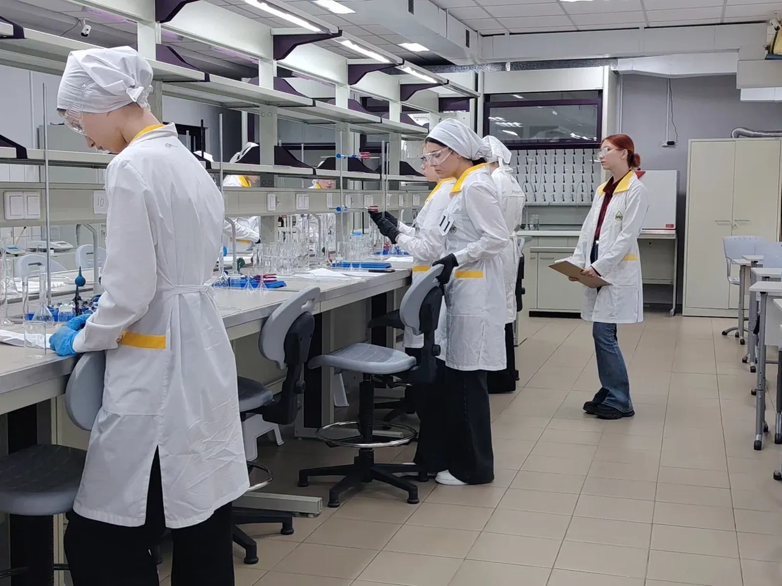 Кадр сделан в современной оборудованной химической или научно-исследовательской лаборатории. За длинными столами-стеллажами с различным стеклянным лабораторным оборудованием (колбы, мензурки) работают несколько человек. Одна из девушек стоит спиной к камере, проводя эксперимент в защитных очках и перчатках. Другая девушка справа стоит прямо, держит в руках планшет для записей, на ней белый лабораторный халат с жёлтой отделкой. В лаборатории светло, пол выложен светлой плиткой.