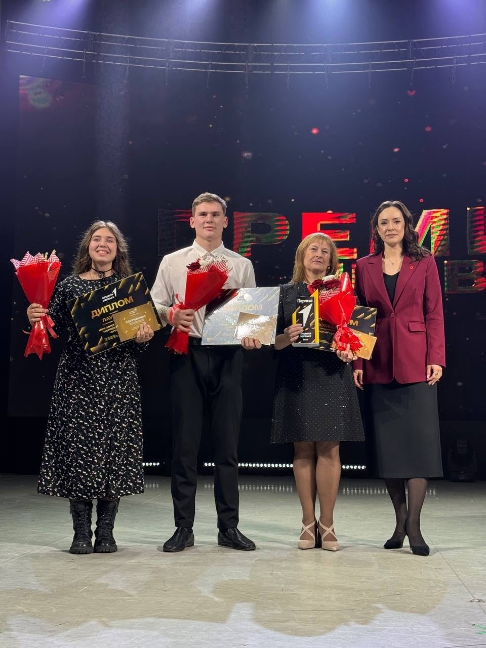 На сцене стоят четыре человека. Слева направо: две женщины, одна из которых в черном платье с белыми точками, а другая в черной юбке и блузке. Они держат в руках дипломы и цветы. В центре изображен молодой человек в белой рубашке, который также держит диплом и цветы. Справа — женщина в темно-синем костюме с длинными волосами, которая улыбается и стоит рядом с награжденными. На заднем плане виден яркий экран с надписью на ломаном шрифте "Премия Первых".