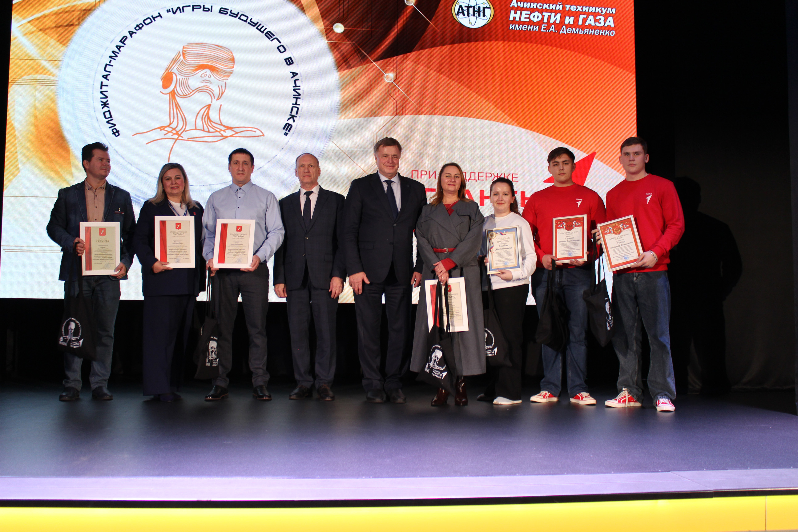 На этой фотографии группа людей стоит на сцене, идущей к зрителям. Они держат дипломы и награды, выражая радость. Посередине среди них – мужчина в строгом костюме, который говорит с ними. Все выглядят счастливыми и гордыми, на заднем плане снова видно яркую проекцию с логотипами и текстом мероприятия, которое они представляют.