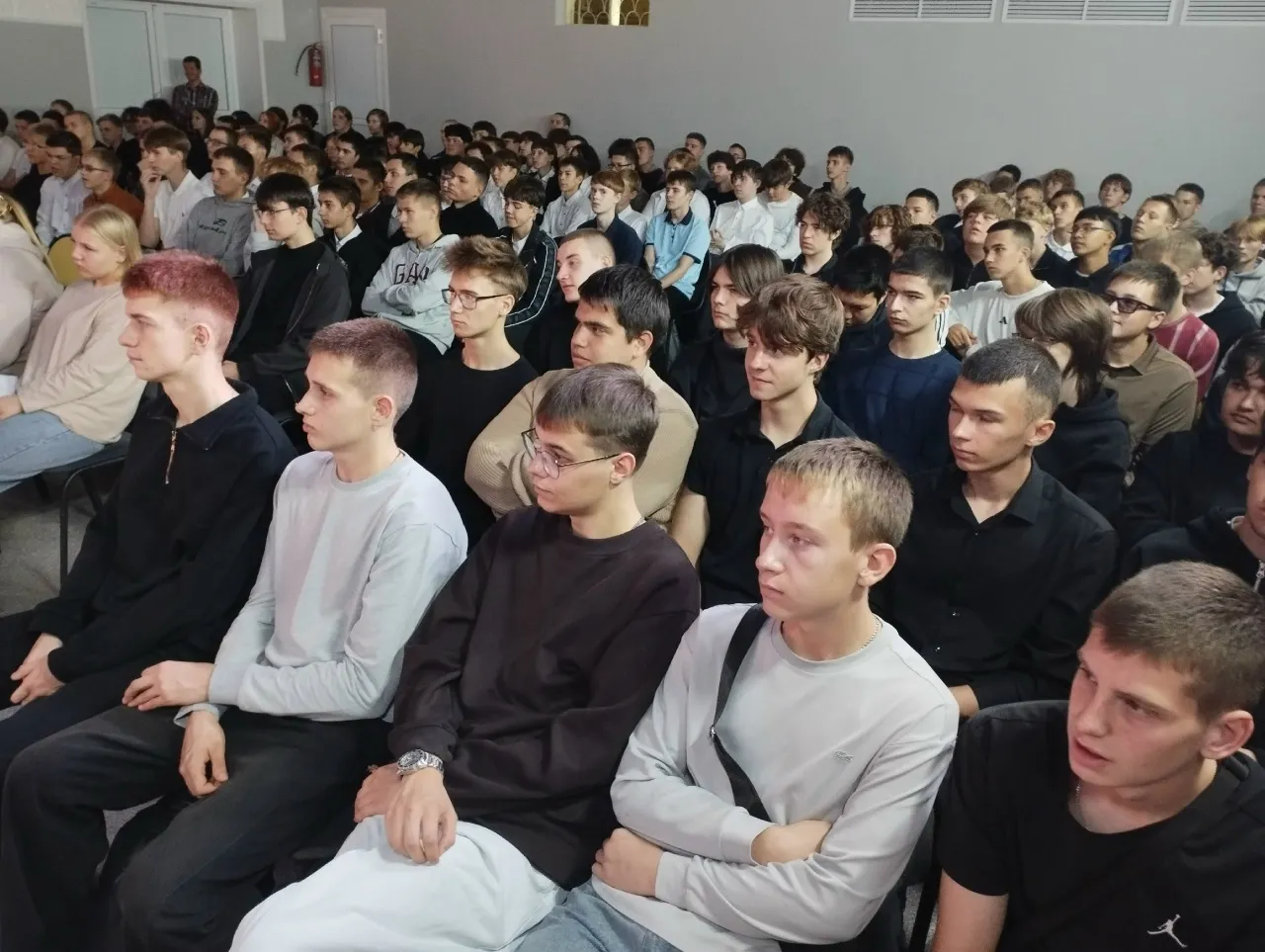 Большая группа молодых людей, преимущественно парней, сидит в зрительном зале и внимательно смотрит влево. На переднем плане выделяются парень с рыжеватыми волосами в черной кофте, парень в светло-сером свитере, парень в очках и темном свитере, а также молодой человек в сером свитере с черной сумкой через плечо. Люди одеты в casual-стиле: черные, серые, белые футболки и кофты. Зал имеет светлые стены, на заднем плане видны двери и огнетушитель на стене.