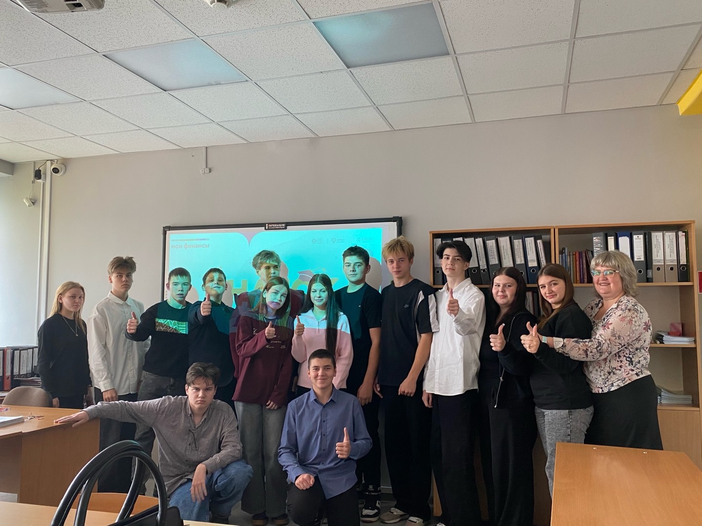 BfNK96kJ0IDUtQF1ws_-3На фотографии видно группу из 15 человек, которые стоят в классе. Позади них находятся папки и шкафы с документацией. Все участники улыбнувшиеся и стоят в разных позах, но большинство из них показывают палец вверх в знак одобрения. Атмосфера дружелюбная и весёлая, стены светлые.ni0Bi46x954c67Q4BUdCgdxB30zW5FWAZyoBiQH8RjRhPff-onhnU6urR8YmNRTn9IR.jpg