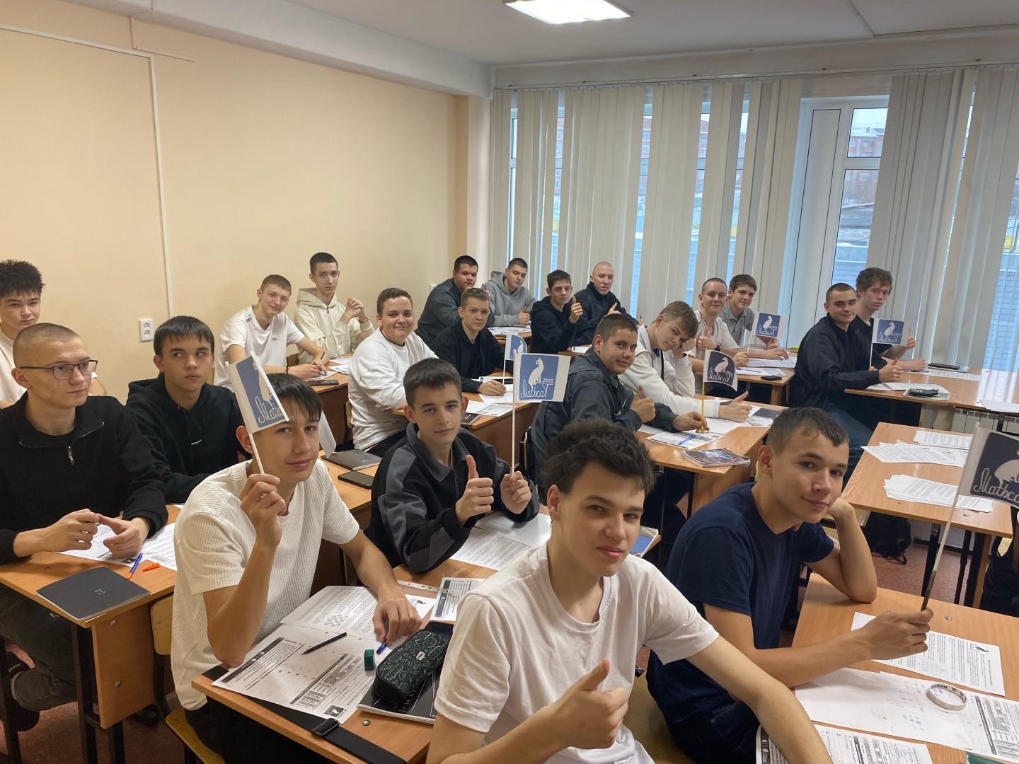 На фотографии изображена группа из семнадцати учеников, сидящих за столами в классной комнате. У каждого из них в руках фотографии или карточки, которые они держат высоко. Большинство из них улыбаются и показывают жест пальцем "ок", что указывает на положительное отношение. Стены комнаты светлые, а окна с вертикальными жалюзи, пропускающими дневной свет.