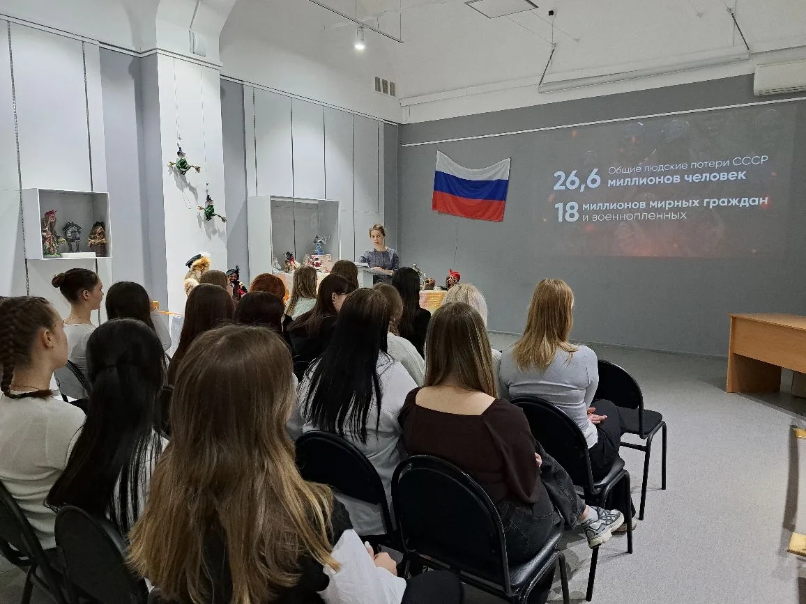 изображён зал, где проводится мероприятие. На стене висит флаг России, а на экране видно текст с цифрами: 26,6 миллиона человек и 18 национальных меньшинств. В центре зала стоит женщина, объясняющая информацию присутствующим, которые внимательны к её словам. Помещение оформлено в светлых тонах, с несколькими предметами декора на стенах.