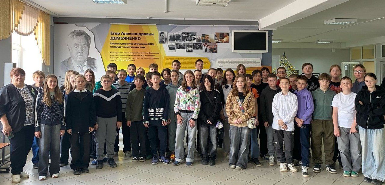 На изображении собрана группа молодых людей и взрослых, стоящих в помещении. Все они стоят в несколько рядов, на заднем плане виден плакат с фотографиями и текстом, который начинает с фразы "Этому Александрову Девяткин". Участники выглядят дружелюбно и заинтересованно, многие из них улыбаются. Стены помещения светлые, с витринами и телевизором на стене.