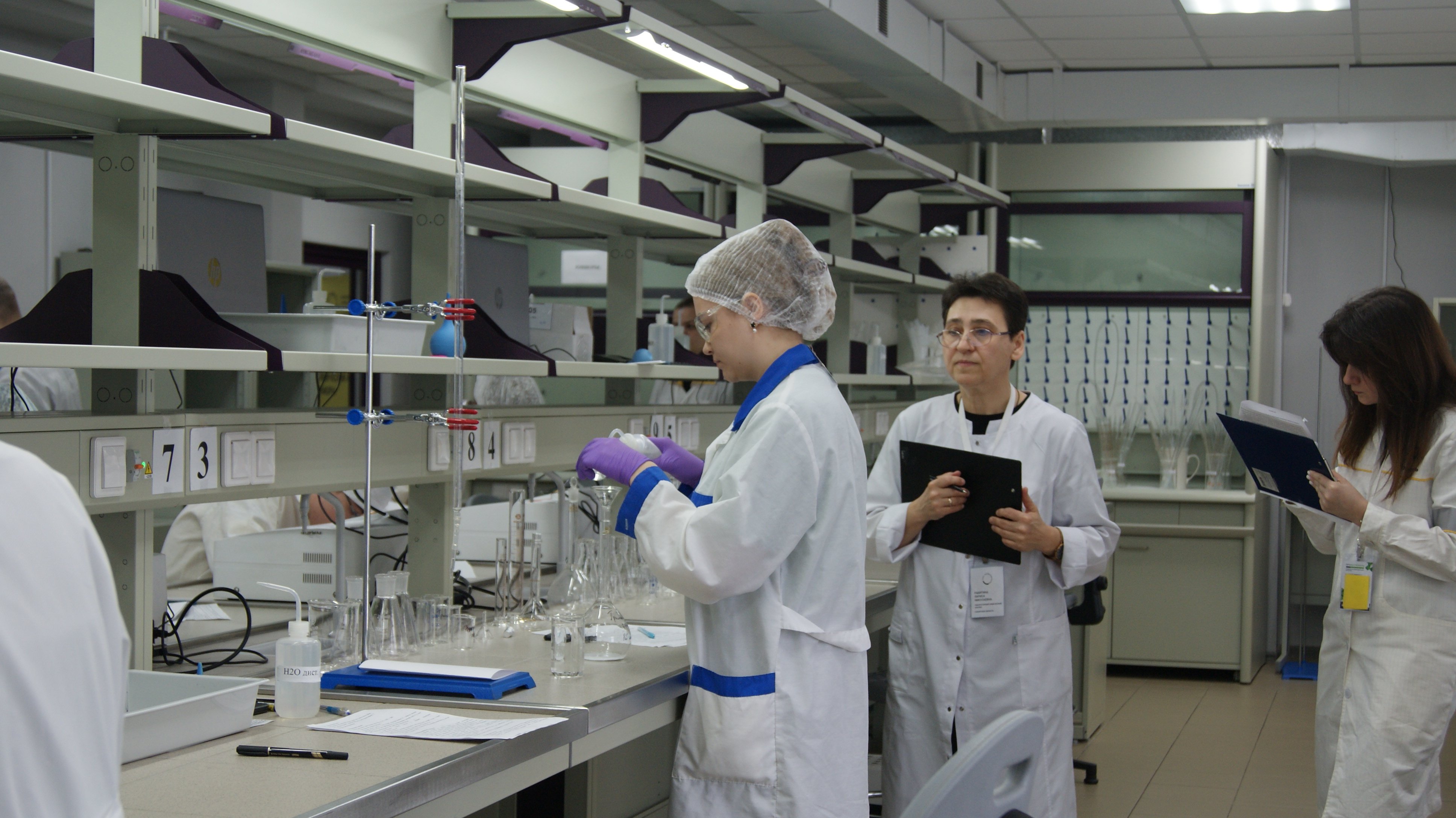 На данном фото видно двух ученых в белых халатах, работающих в лаборатории. Один из них, женщина, измеряет жидкости в пробирках с помощью пипетки, а другой, мужчина, наблюдает и записывает данные. Все вокруг них обставлено современным оборудованием и лабораторными принадлежностями. Место хорошо освещено.