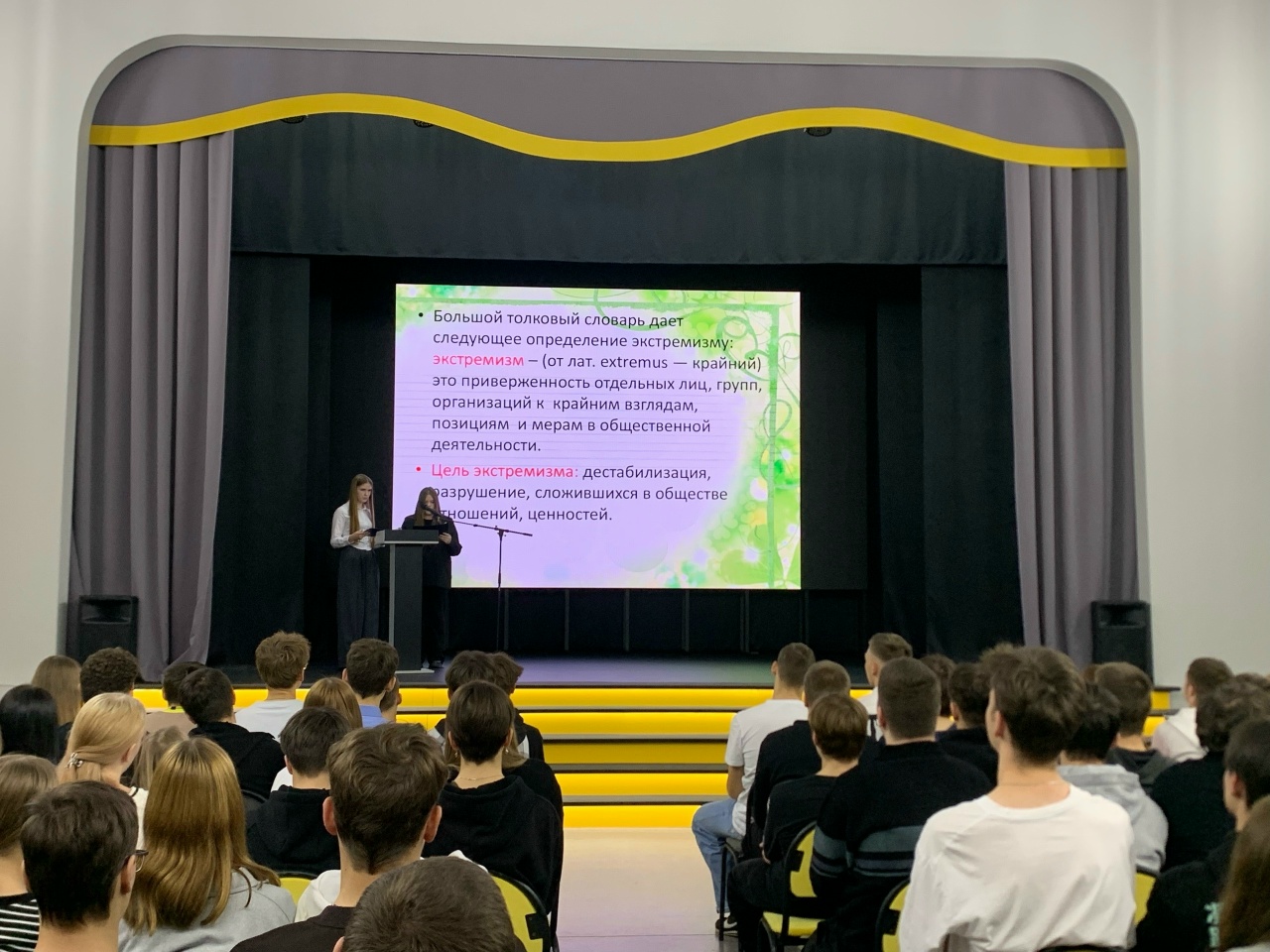 Женщина продолжает свою презентацию. На экране виден текст с длинным описанием, относящимся к темам экстремизма и терроризма. Аудитория сосредоточена на спикере, многие колонны стульев заняты молодежью в темной одежде.