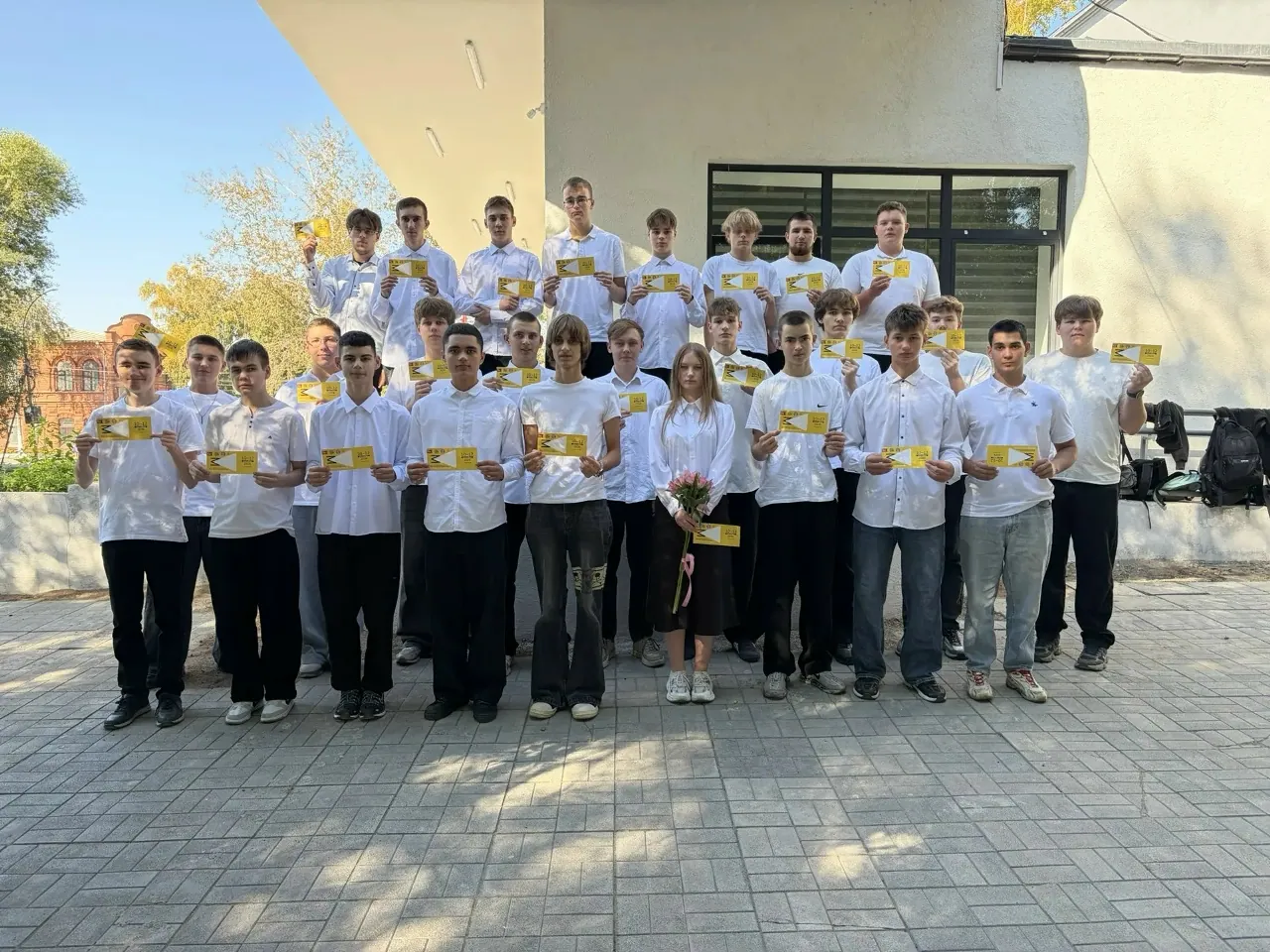Групповое фото молодых людей и мужчины на улице у здания в солнечный день. Большинство одеты в белые рубашки или футболки и темные брюки. Почти все держат в руках желтые карточки или сертификаты. В первом ряду стоит девушка с букетом цветов. На заднем плане видны деревья, окна здания и рюкзаки на скамейке.