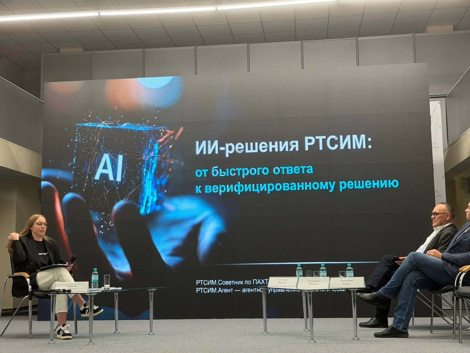 Три спикера сидят на сцене перед большим экраном. На экране крупная надпись «ИИ-решения РТСИМ: от быстрого ответа к верифицированному решению» и изображение руки, держащей светящийся синий куб с надписью «AI».