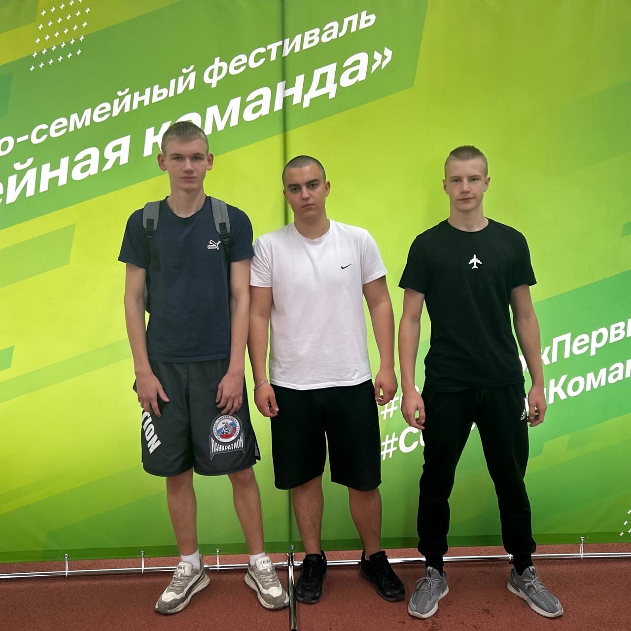 На изображении три молодых человека стоят на фоне яркого баннера с надписью о фестивале. Все они одеты в спортивную одежду: двое в футболках с короткими рукавами, один — в черной футболке. Они выглядят уверенно и улыбаются, позируя для фотографии. Фон баннера яркий, с геометрическими узорами и текстом.