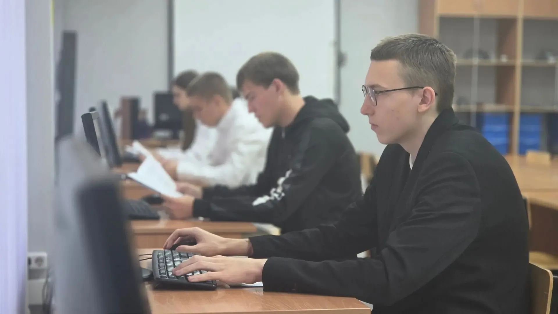 Молодой человек в очках и черной одежде сидит за деревянным столом и печатает на клавиатуре, глядя на экран монитора. Он находится в компьютерном классе, позади него в ряд сидят другие студенты, один в черной толстовке и один в белой рубашке, которые также работают за компьютерами. На заднем плане виден деревянный стеллаж.