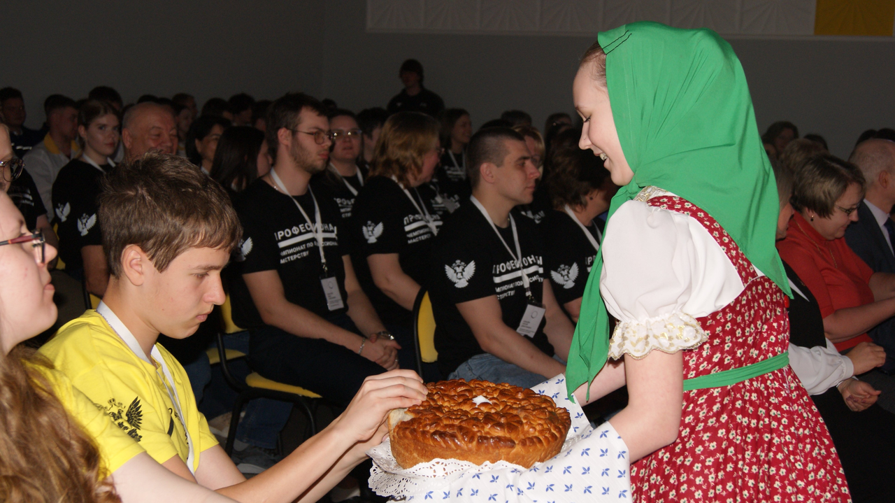 На этом изображении молодая девушка в традиционном костюме с зеленым платком на голове предлагает мужчине с короткой стрижкой хлеб, украшенный узором. Оба они находятся в зале, полном зрителей, которые внимательно смотрят на происходящее. Атмосфера дружеская и теплое взаимодействие между поколениями подчеркивает культурные традиции.